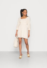 Vestido mini floral em amarelo claro e branco com mangas bufantes, decote quadrado e corte solto, combinado com sandálias amarelas e uma bolsa bege.