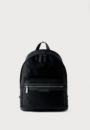 Sac à dos noir avec poche zippée à l'avant et double curseur de fermeture éclair sur le dessus, posé debout sur un fond gris clair uni.