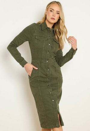 Pitkähiuksinen nainen, joka on pukeutunut istuvaan oliivinvihreään, nappikiinnitteiseen midimekkoon, jossa on pitkät hihat ja sivutaskut.