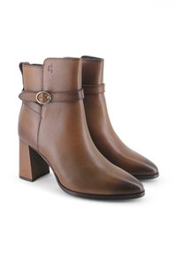 Botas de cuero marrón hasta el tobillo con punta afilada, tacón cuadrado y una correa decorativa con una hebilla circular. Textura suave, diseño simple.