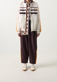 Lätt, off-white bomberjacka med bruna ränder, ihop med mörkbruna, löst sittande byxor med orange sidostripes och vita sneakers.