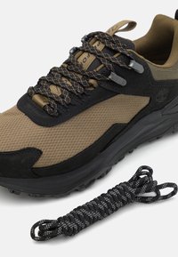 Bruine en zwarte sneaker met een bovenwerk van textured mesh, suede accenten, een dikke zwarte rubberen zool en zwart-grijze gemusterde veters ernaast.