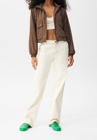 Brązowa krótka kurtka z wysokim kołnierzem, biały krótki top, beżowe spodnie o szerokich nogawkach i zielone masywne buty. Gładka faktura, minimalistyczne detale.