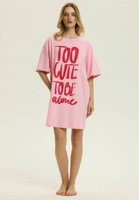 Młoda kobieta stoi boso, ubrana w luźną różową sukienkę z czerwonym napisem „TOO CUTE TO BE alone” na jednolitym tle.