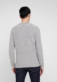 Helles graues Langarmpullover mit strukturiertem Strickmuster, Rundhalsausschnitt und einer leicht taillierten Form, kombiniert mit dunklen Jeans.