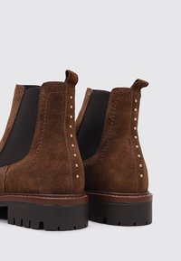 Braune Wildleder-Stiefeletten mit elastischen Seitenpaneelen, runder Zehenpartie und dekorativen goldenen Nieten entlang der Rückennaht. Schwarze Gummisohle und -absatz.