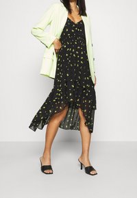 Frau in einem schwarzen, floralen High-Low-Kleid, kombiniert mit einem hellgrünen Blazer und schwarzen, offenen High-Heel-Mules, die vor einem weißen Hintergrund steht.