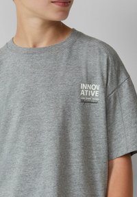 Grijze katoenen t-shirt met korte mouwen, voorzien van een witte bedrukte logo "INNOVATIEF" en aanvullende tekst. Zachte textuur en relaxte pasvorm.