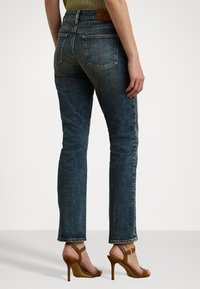 Donkerblauwe denim jeans met een taps toelopende pijp, voorzien van achterzakken en een leren label op de tailleband, gecombineerd met bruin hoge hakken.