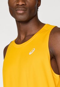 Κίτρινο αθλητικό τοπ με στρογγυλή λαιμόκοψη, που διαθέτει ένα μικρό λευκό λογότυπο στο στήθος. Το ύ φασμα είναι υφασμένο και ελαφρύ.