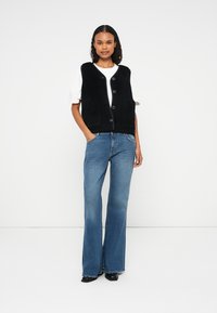 Black button-up vest made of textured fabric, worn over a white shirt, paired with light blue flared jeans and black ankle boots.