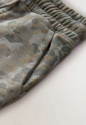 Detailné záber na nohavice so vzorom maskáča, s elastickým pásom a švami bočných vreciek na svetlom pozadí.