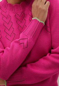 Fuchsia knit sweater with openwork heart patterns, ribbed collar and cuffs edged in light green, worn with one hand resting on chest.