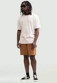 Vīrietis ar dreads, valkā melnus saulesbrilles, baltu T-kreklu, brūnus šortus, melnas zeķes un melnus sporta apavus, stāvot pret vienkāršu fona.