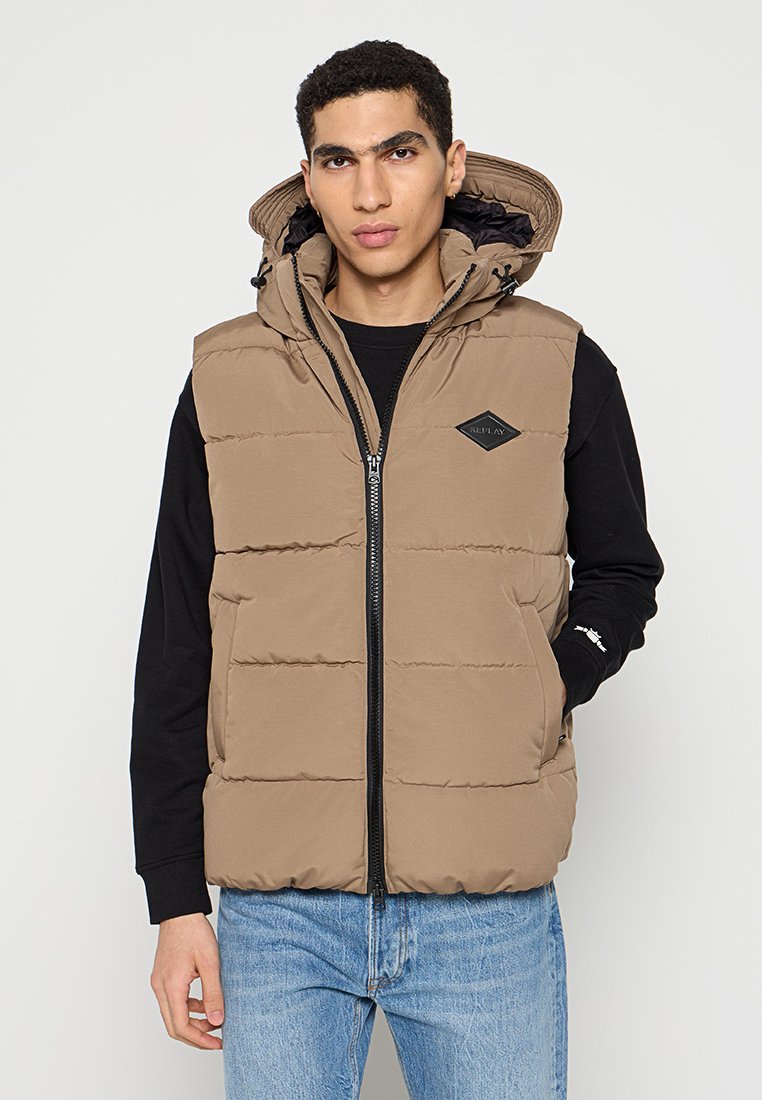 Braune Pufferweste mit Kapuze, Reißverschluss und Seitentaschen. Design mit Panelen und einem Diamantlogo auf der Brust; getragen über einem schwarzen Shirt.