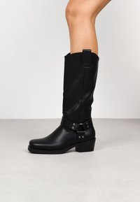 Bota de cuero negro hasta la rodilla con punta cuadrada, superficie texturizada, detalle de hebilla de metal y tacón bajo.