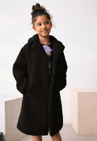 Manteau noir et duveteux avec un col, présentant une texture douce et des manches longues. Porté sur un haut à paillettes, avec une coupe décontractée.