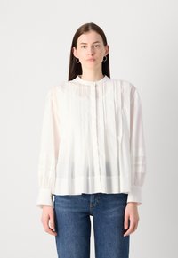 White blouse with pleats, round neckline, and long sleeves. Material is lightweight with a slight sheen. Worn with blue jeans.