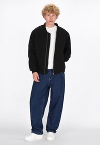 Giacca bomber nera con zip e polsini a coste, abbinata a jeans scuri a gamba larga e sneakers bianche, con una semplice t-shirt bianca sotto.