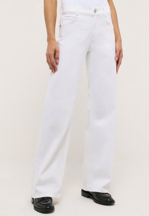 Frau trägt weiße Jeans mit hohem Bund und weitem Bein und schwarze Loafers, steht vor einem schlichten hellen Hintergrund.