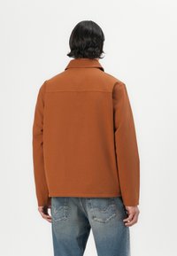 Bruine katoenen jas met een kraag, lange mouwen en een recht model. Gecombineerd met lichtblauwe spijkerbroeken met stikseldetails.