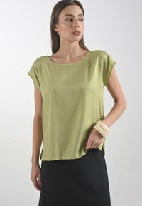 Tričko s krátkým rukávem ve světle zelené barvě s volným střihem. Má kulatý výstřih a postranní rozparky, v kombinaci s černou sukní a slonovinovým náramkem.