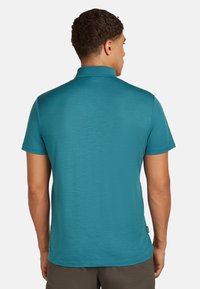 Turkos t-shirt med kort ärm och klassisk krage, texturerad tyg och rak nederkant, med en liten logotypetikett på sidan.