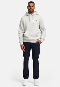 Maglione grigio chiaro con tasca frontale, coulisse e piccolo logo. Abbinato a pantaloni blu navy e sneakers bianche su uno sfondo neutro.
