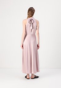 Lång, ärmlös klänning i mjuk rosa tyg med halterneck och en knytbåge i ryggen, flödande ner till anklarna. Slät textur.