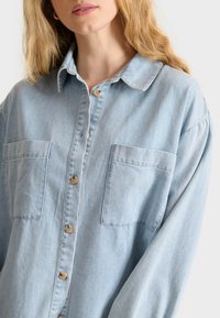 Lichtblauw denim overhemd met een kraag, twee voorzakken en tan kleurige knopen, met een ontspannen pasvorm en een zachte textuur.