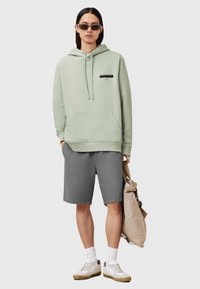 Felpa verde chiaro con cappuccio e coulisse, tasca a marsupio e grafica nera. Pantaloni corti grigi, calzini bianchi e sneakers beige. Porta una borsa beige.