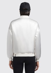 Giacca bomber bianca in satin con polsini e orlo a costine, caratterizzata da una vestibilità comoda. Indossata con un cappellino nero, mostrando una texture liscia e lucida.