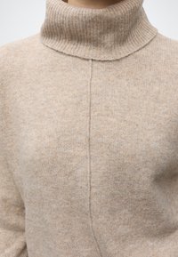 Jersey de cuello alto beige de punto con una costura central visible en el pecho, llevado por una persona sobre un fondo liso.