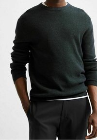 Man med en mörk strukturerad långärmad tröja över en vit skjorta, med svarta byxor och händerna delvis i fickorna.