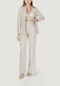 Completo beige con giacca su misura e pantaloni a palazzo a vita alta. Sotto, un reggiseno in pizzo bianco. Tessuto liscio e dettagli con bottoni lucidi.