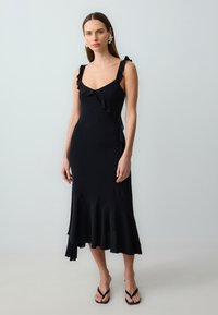 Schwarzes Kleid mit Rüschenträgern, einem taillierten Oberteil und einem ausgestellten Saum. Mittel-lang, glatte Textur und minimale Details.