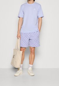 Světlé fialové tričko s malým logem, vzorované světlé fialové šortky, bílé tenisky a béžová taška přes rameno s textem.