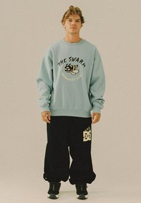 Man staat in een lichtblauwe sweatshirt met een bijenprint en de tekst "The Swarm," zwarte baggy broek met logo, en zwarte sneakers.