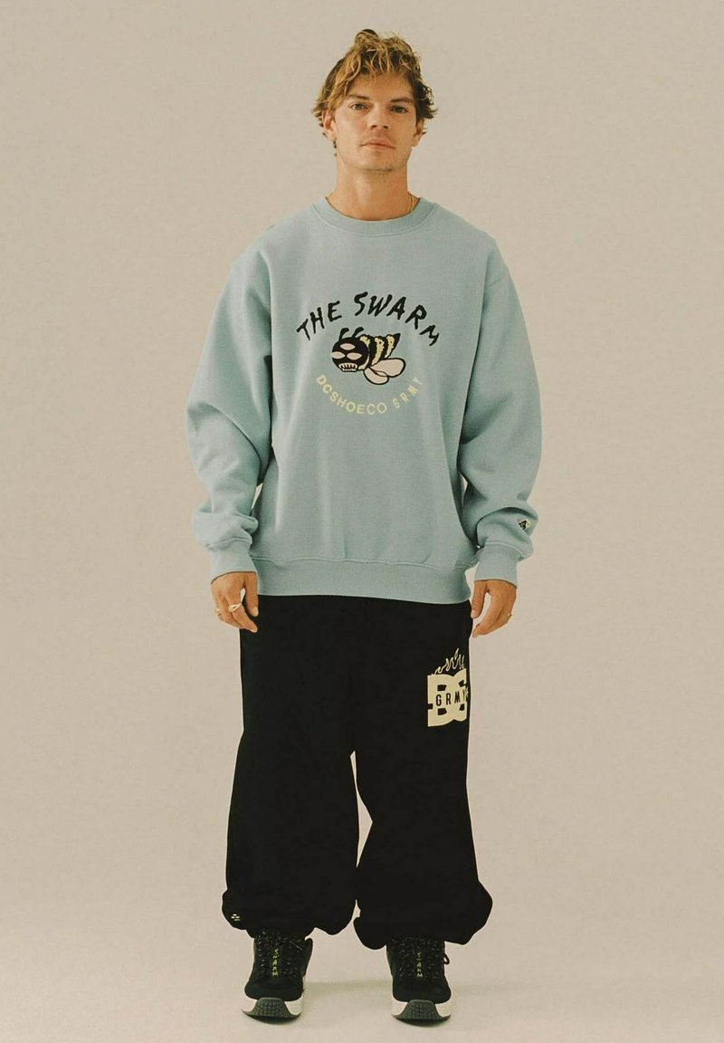 Man staat in een lichtblauwe sweatshirt met een bijenprint en de tekst "The Swarm," zwarte baggy broek met logo, en zwarte sneakers.