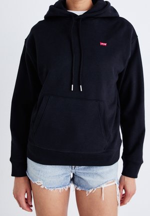 Personne portant un sweat à capuche noir Levi's avec des cordons et un short en jean bleu clair délavé sur un fond uni.