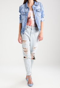 Giacca di jeans azzurro chiaro con dettagli strappati, t-shirt bianca con grafica di un fenicottero, jeans chiari strappati e tacchi blu con decorazioni.