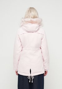 Mulher vestindo um parka rosa claro com capuz forrado a pelo e cordão no hem, vista de costas contra um fundo branco liso.