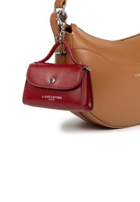 Piccolo astuccio in pelle rossa di Lancaster Paris attaccato a una borsa in pelle beige con hardware argentato su uno sfondo bianco.