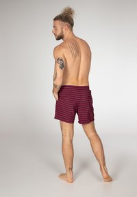 Bordeaux kleurige zwemshorts met dunne witte strepen, die een relaxte pasvorm en een elastische tailleband laten zien. Het model staat op blote voeten met de rug naar de camera gekeerd.