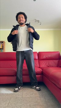 Schwarze Pufferjacke über einem weißen T-Shirt, kombiniert mit dunklen Jeans und schwarzen Sneakers mit weißen Akzenten. Rotes Sofa im Hintergrund.