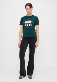 T-shirt de algodão verde com estampado "Feliz Natal" e gráficos de pinguins, combinada com calças flare pretas brilhantes.