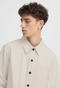 Chemise beige avec une texture douce, dotée d'un col large et de boutons noirs sur le devant. Le tissu semble léger et décontracté.