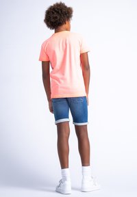 Camiseta rosa brillante, de manga corta, y pantalones cortos de mezclilla azules con dobladillos enrollados. Zapatillas blancas y calcetines blancos. Vista trasera, fondo liso.