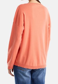 Korálový pletený svetr s uvolněným střihem, spadlými rameny, žebrovaným lemem a jemnou texturou. V kombinaci s modrými denimovými džíny.