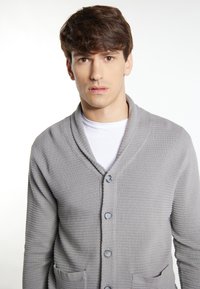 Šedý strukturovaný cardigan s chráničem krku, předními knoflíky a dvěma kapsami. Nosený přes bílou košili s ribovaným designem materiálu.